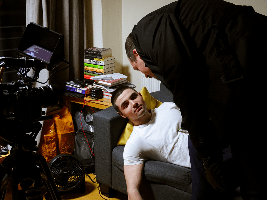 Two actors filming a take as one lies on couch while the other towers over him.