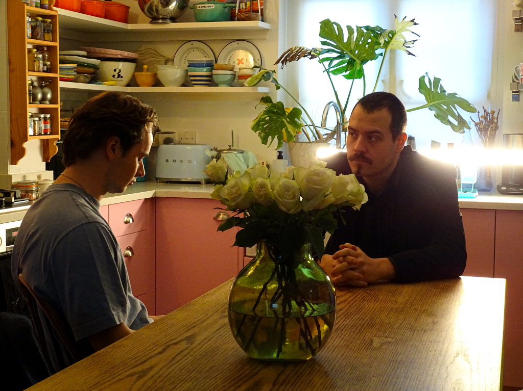 Two actors sitting at table opposite each other.